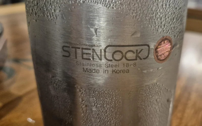 Close-up of a stainless steel object with a logo and text, water droplets on surface.