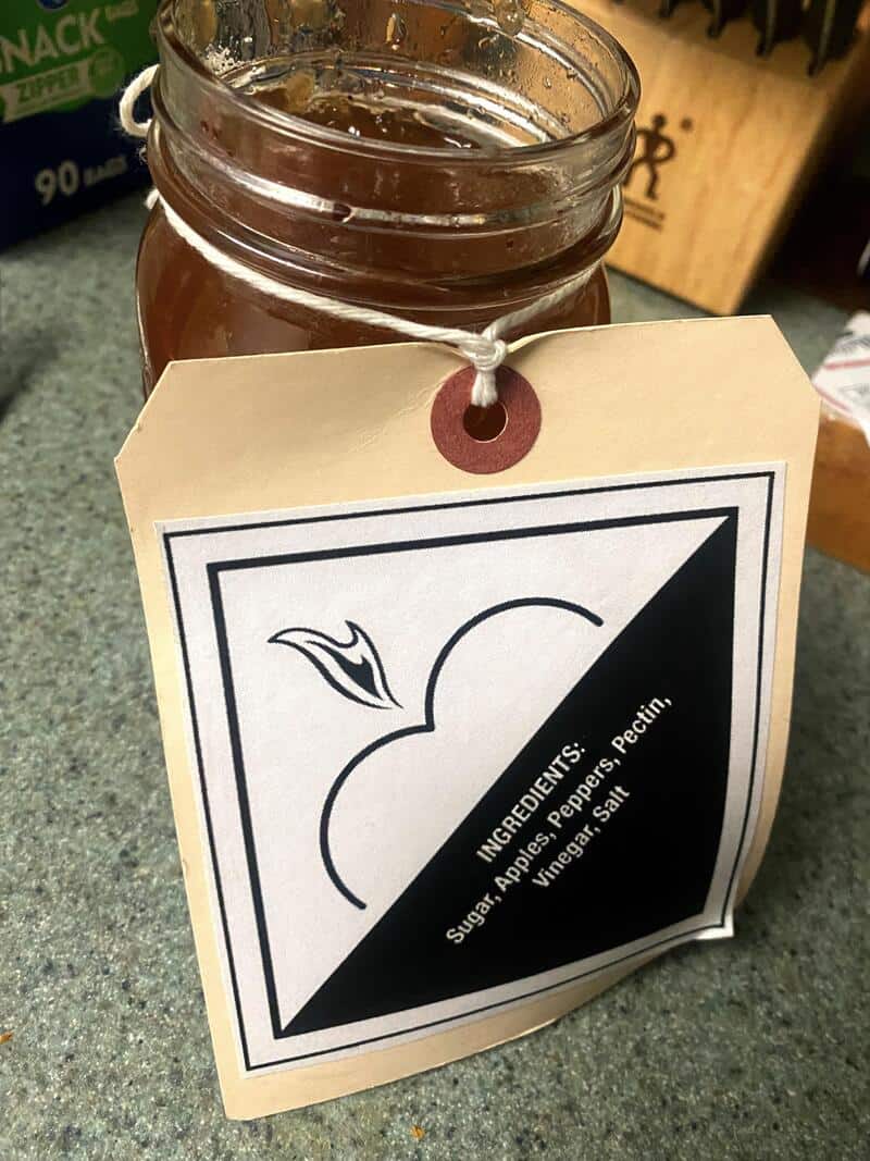 Jar of apple pepper jelly with a label showing ingredients and a black and white apple illustration.