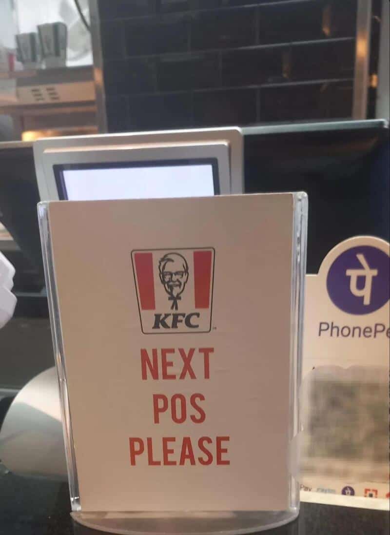 Sign says 'NEXT POS PLEASE' with KFC logo, at a restaurant counter.