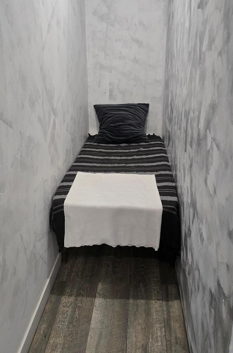 Small bed with black and white striped bedding and a black pillow in a narrow room.