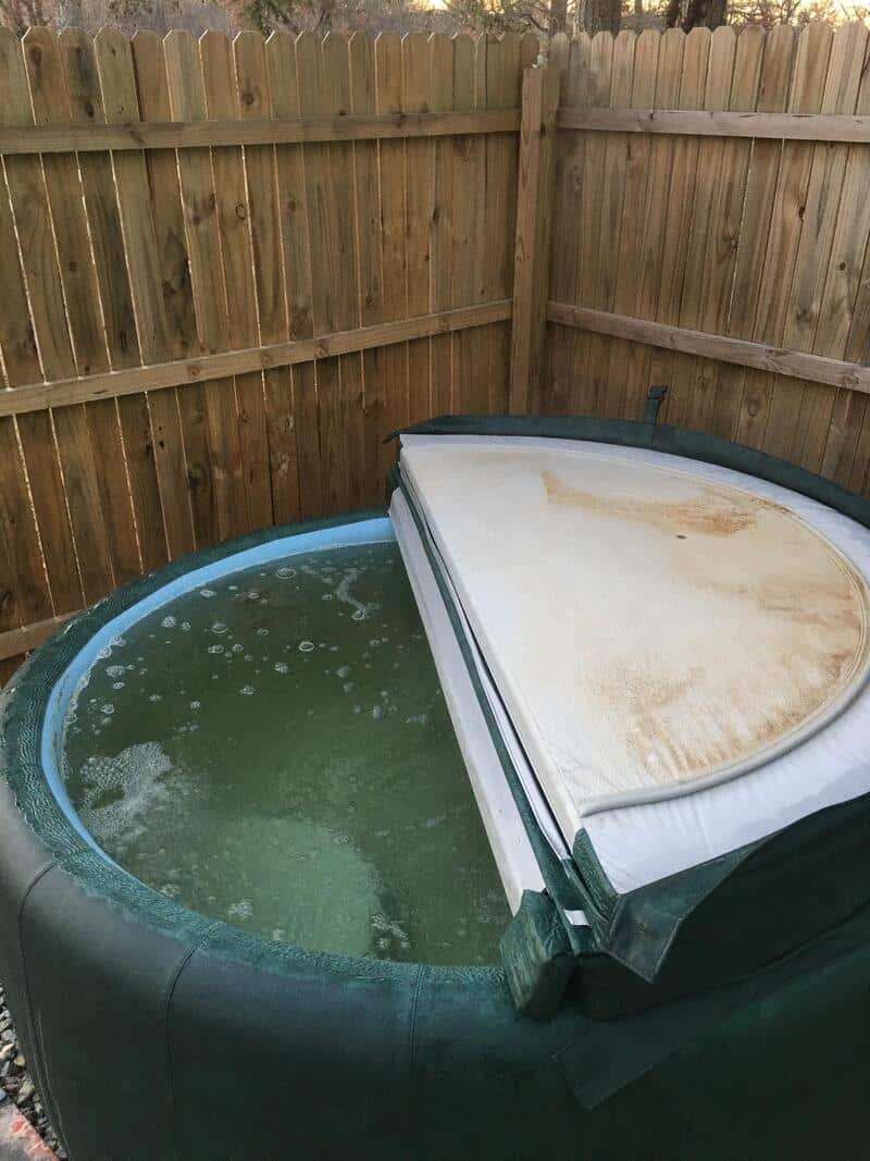 Hot tub with a cover that is half open, filled with greenish water, in a backyard.