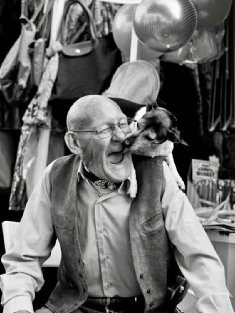 Elderly man with glasses and a dog in a shop, both smiling.