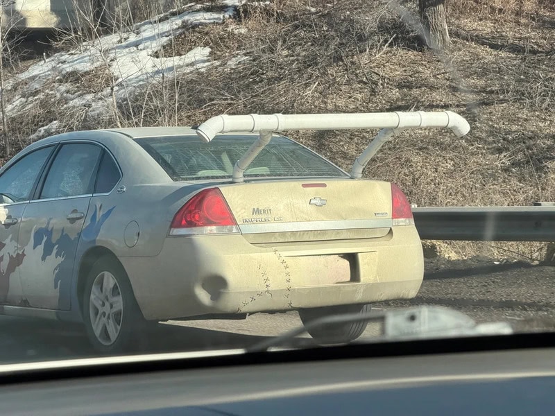 Dirty silver Chevrolet Malibu with a large white pipe on the trunk.