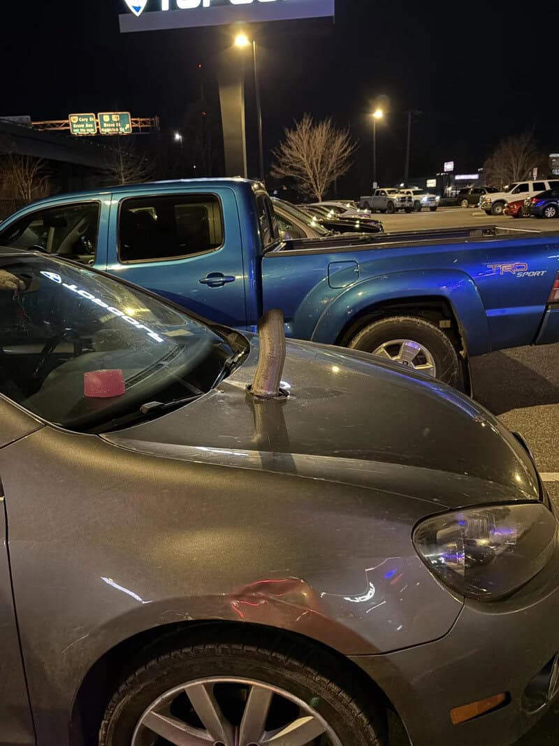 A parking lot at night with a blue pickup truck and a gray car with a dildo on its hood.