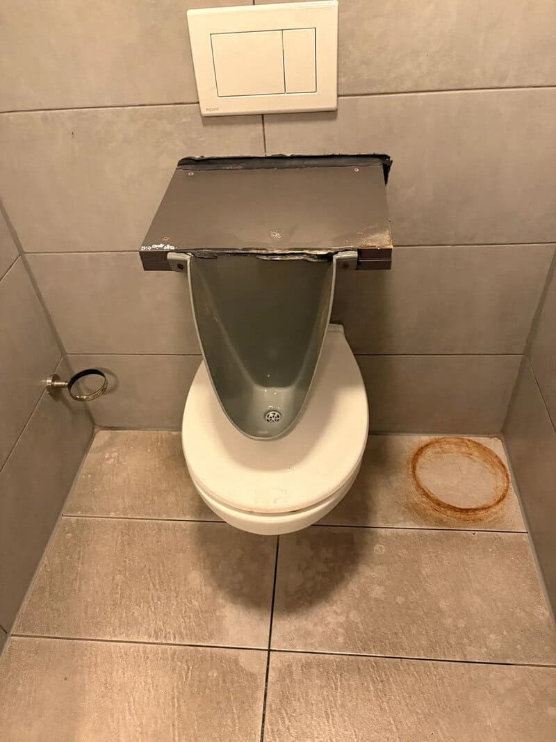 Toilet with a makeshift wooden cover and a rusty floor stain.