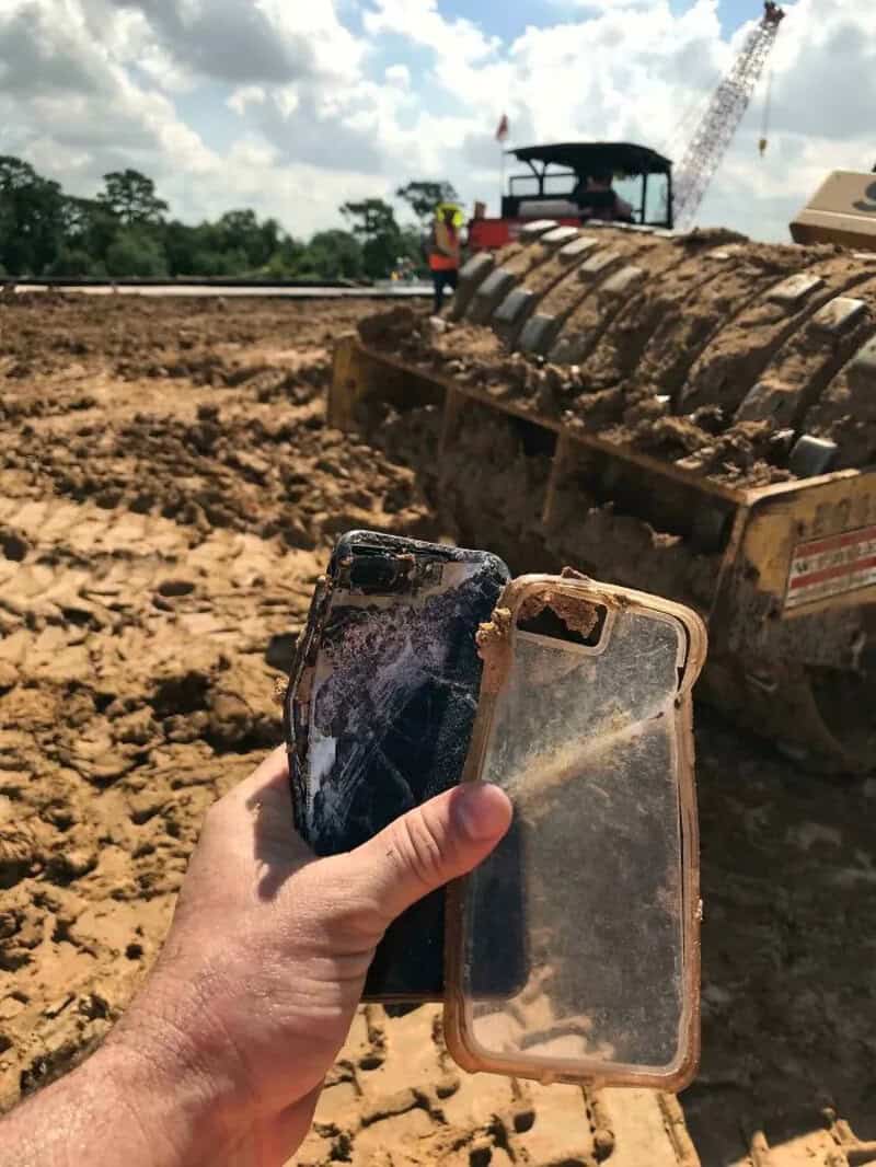 Person holding two damaged smartphones at a construction site with heavy machinery in background.