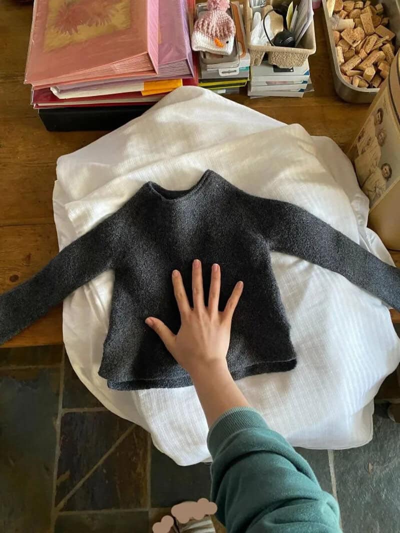 A small black sweater with long sleeves laid out on a white cloth, with a hand touching it.