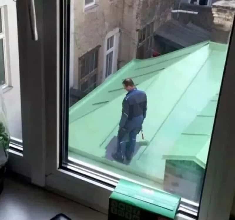 Man on green roof seen through window, holding a tool, with old buildings in background.