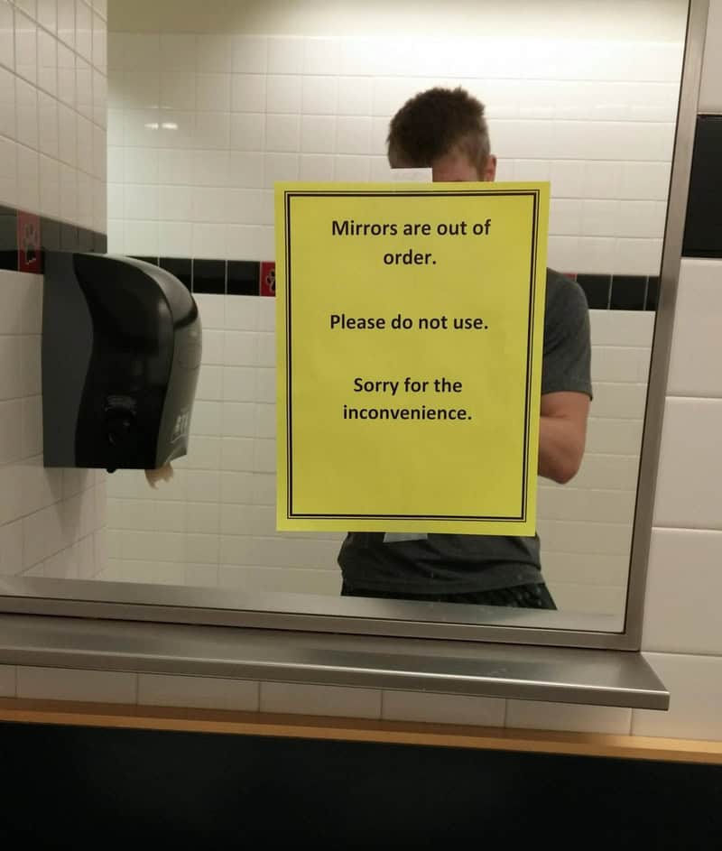 Mirror with a yellow sign that says mirrors are out of order, please do not use, sorry for the incon.