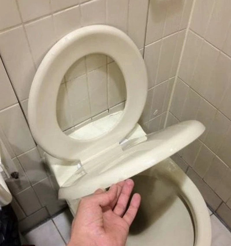 Toilet with a hand pointing at the seat, in a tiled bathroom.