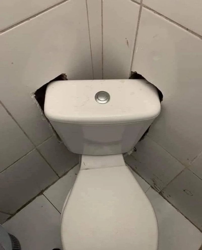Toilet with a hole in the wall behind it, in a bathroom.