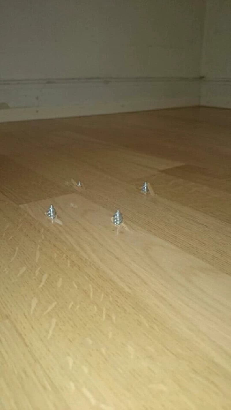 Four screws on a wooden floor near a wall, part of a furniture assembly.