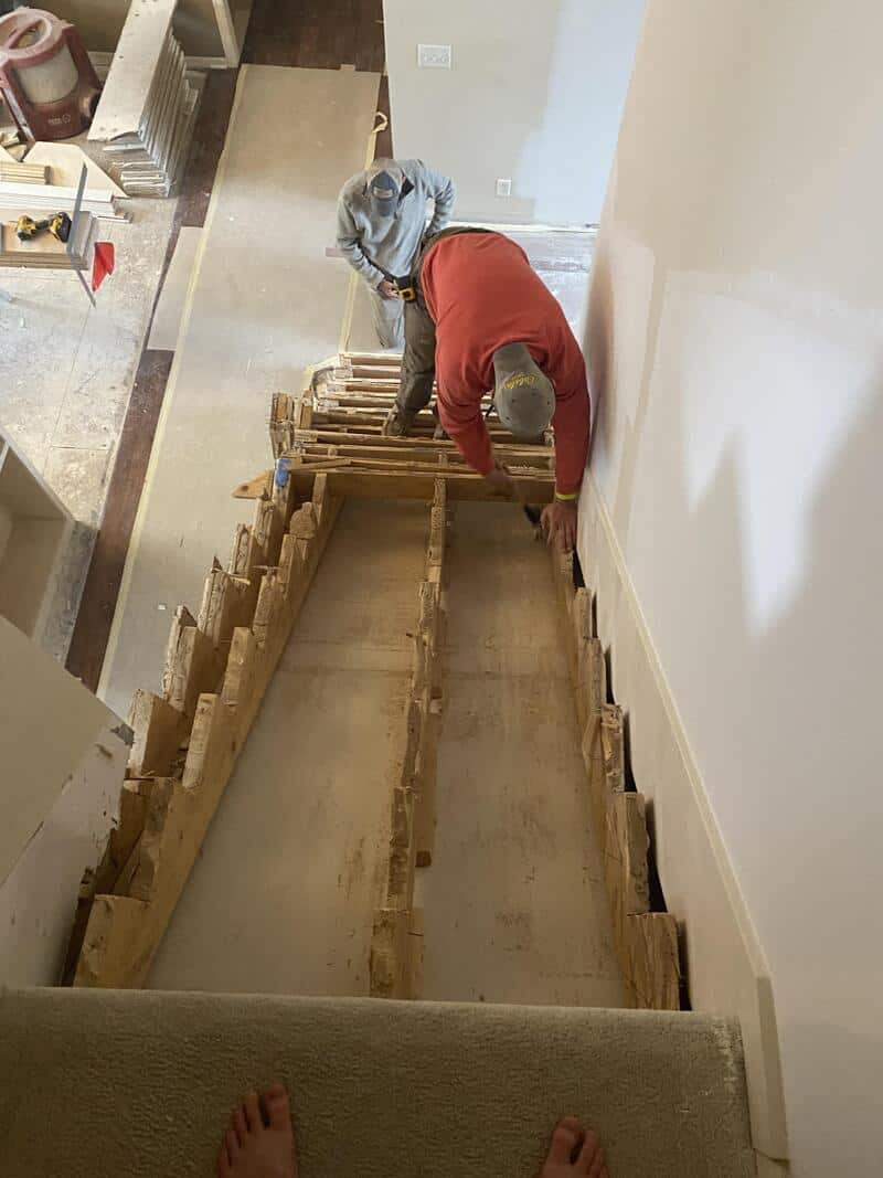 Two workers repair a staircase, one leaning over the railing, the other working on the steps.