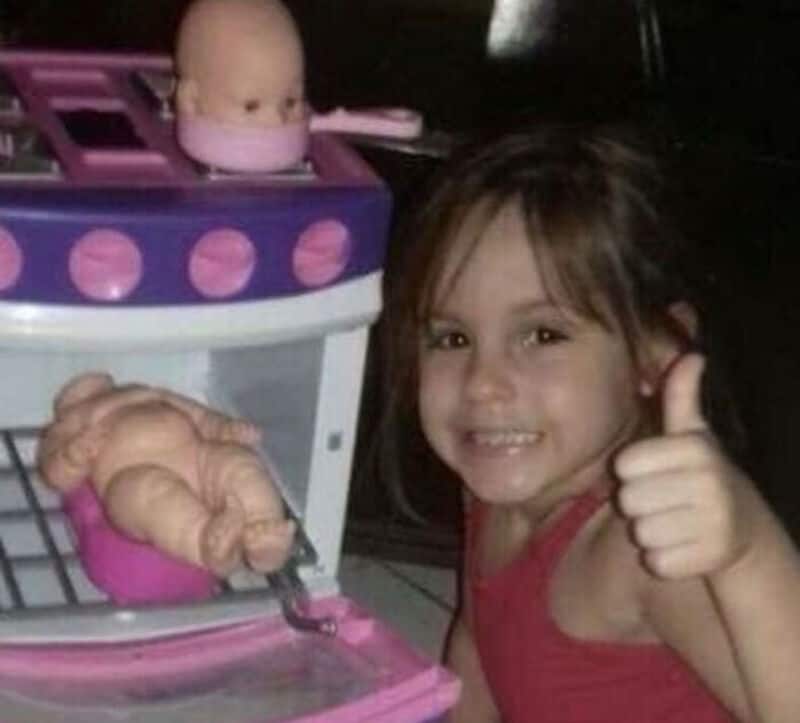 Girl giving a thumbs up next to a toy kitchen with a baby doll and toy food.