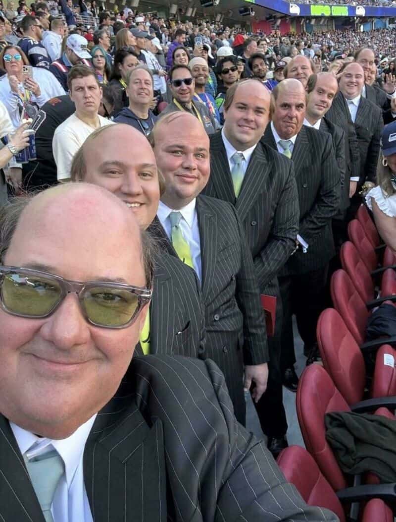 Group of men in suits at a stadium, smiling for a selfie.
