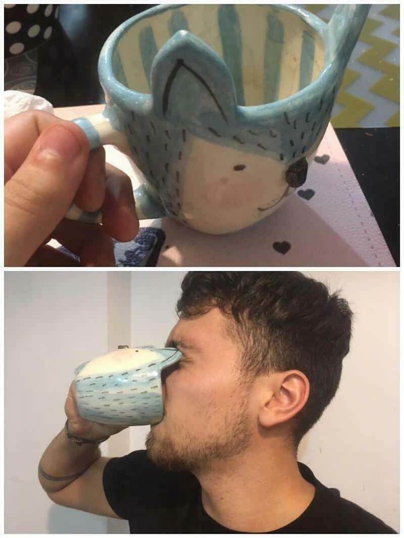 Person drinking from a mug with a face and a hat design, mug held with hand.