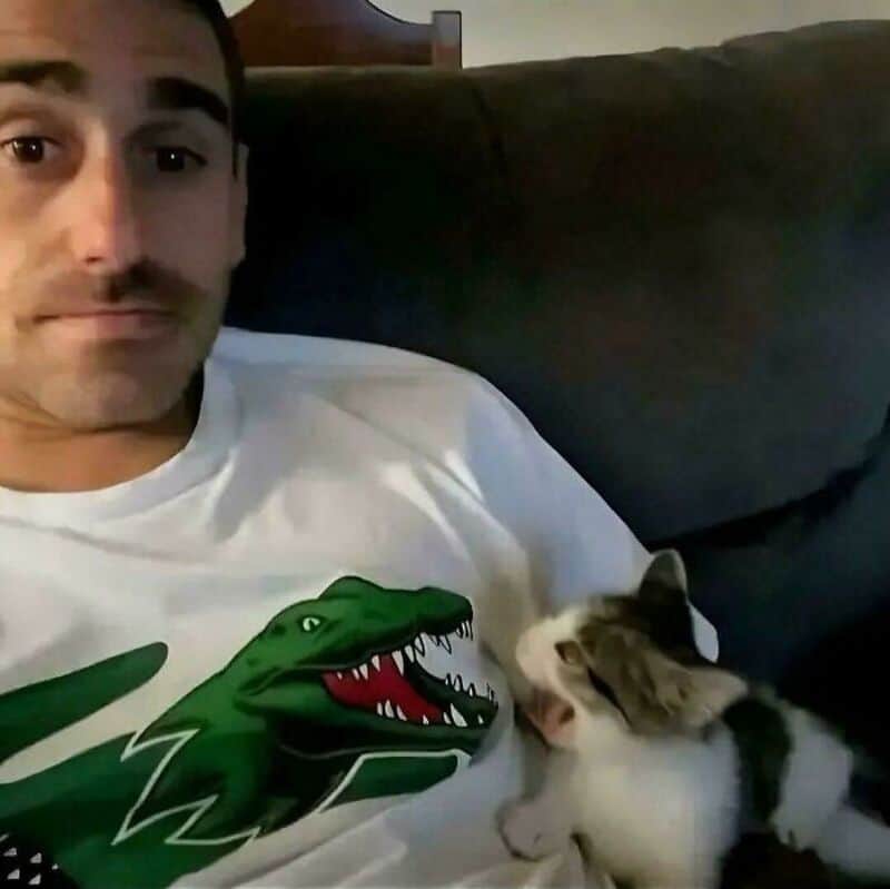 Man in a white T-shirt with a green crocodile logo, sitting on a couch with a cat.