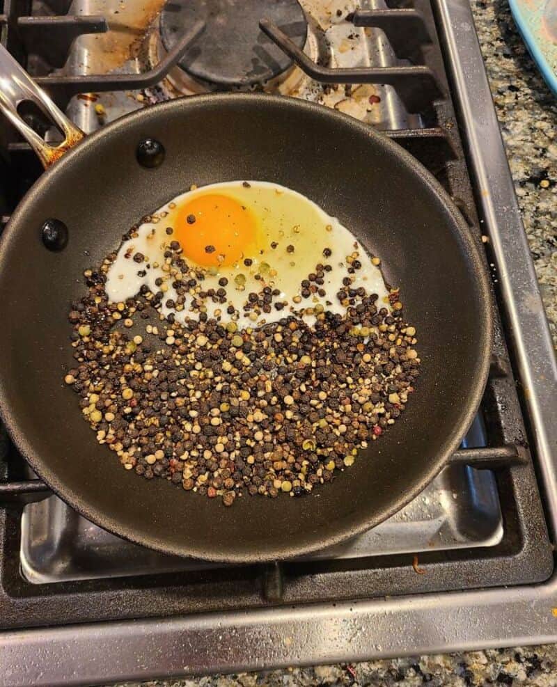 Fried egg with black pepper and mustard seeds in a frying pan on stove.