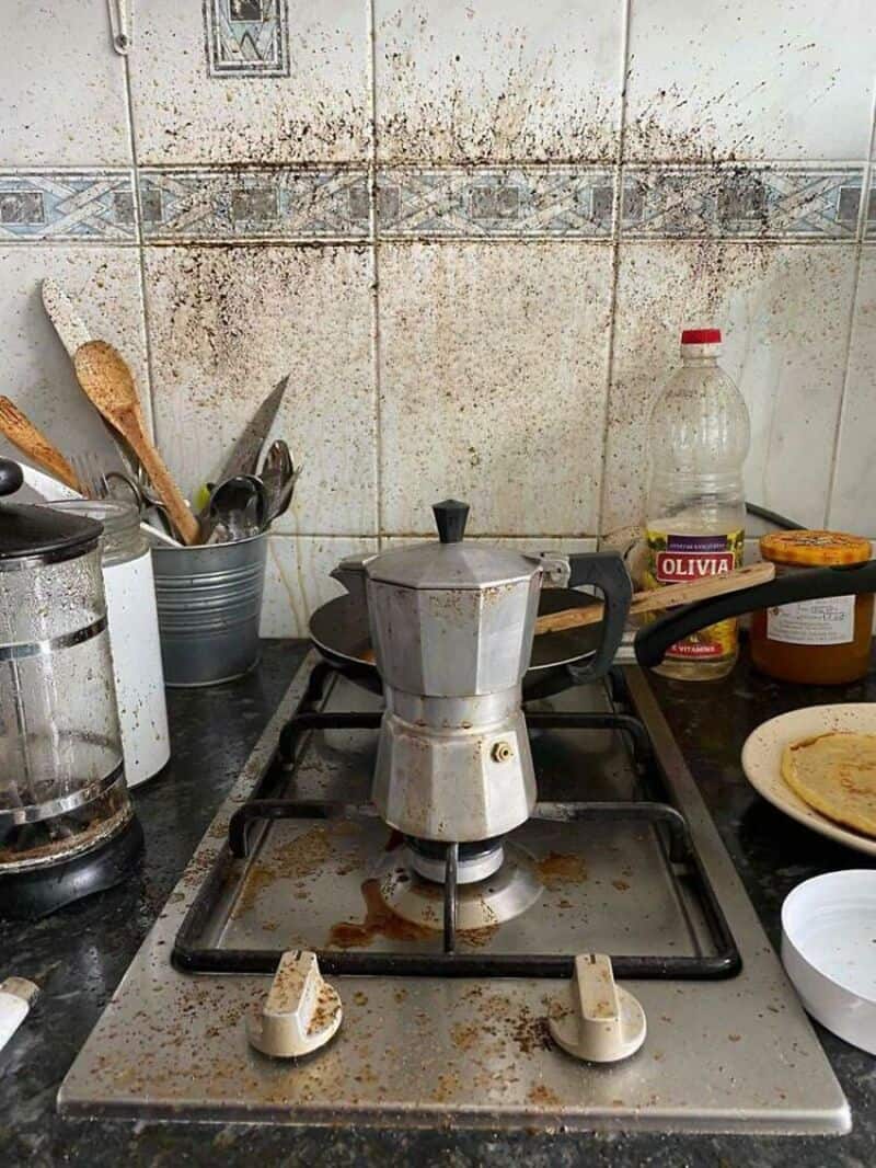 Dirty stove with a coffee maker and kitchen utensils, stained wall behind.