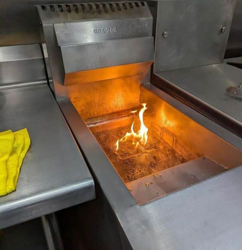 Kitchen deep fryer with fire inside, under a metal hood, with a yellow cloth nearby.
