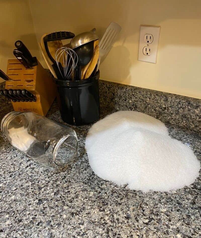 Spilled sugar on kitchen counter with utensils and glass jar nearby.