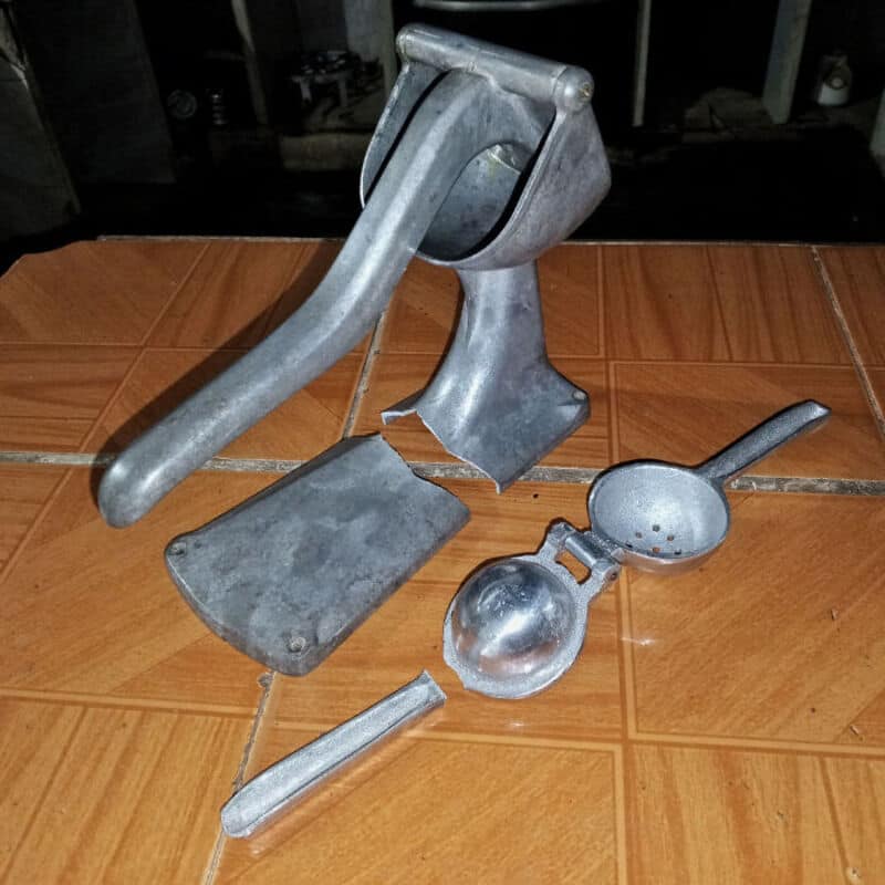 Broken metal kitchen tools on a tiled floor, including a meat tenderizer and a garlic press.