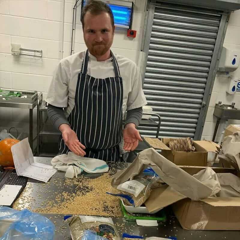 Man in kitchen with spilled grains and open boxes, wearing apron and smiling.