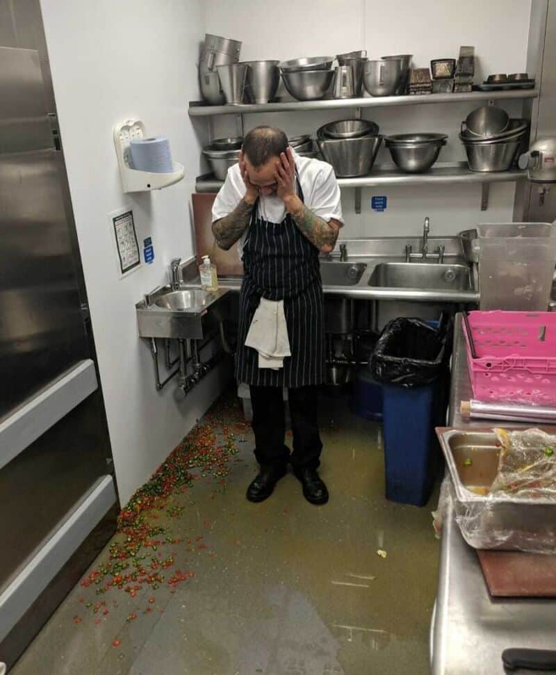 Chef with head in hands in a messy kitchen with spilled food on the floor.