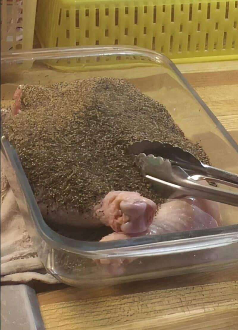 Raw chicken in a glass dish with black pepper and tongs on top.