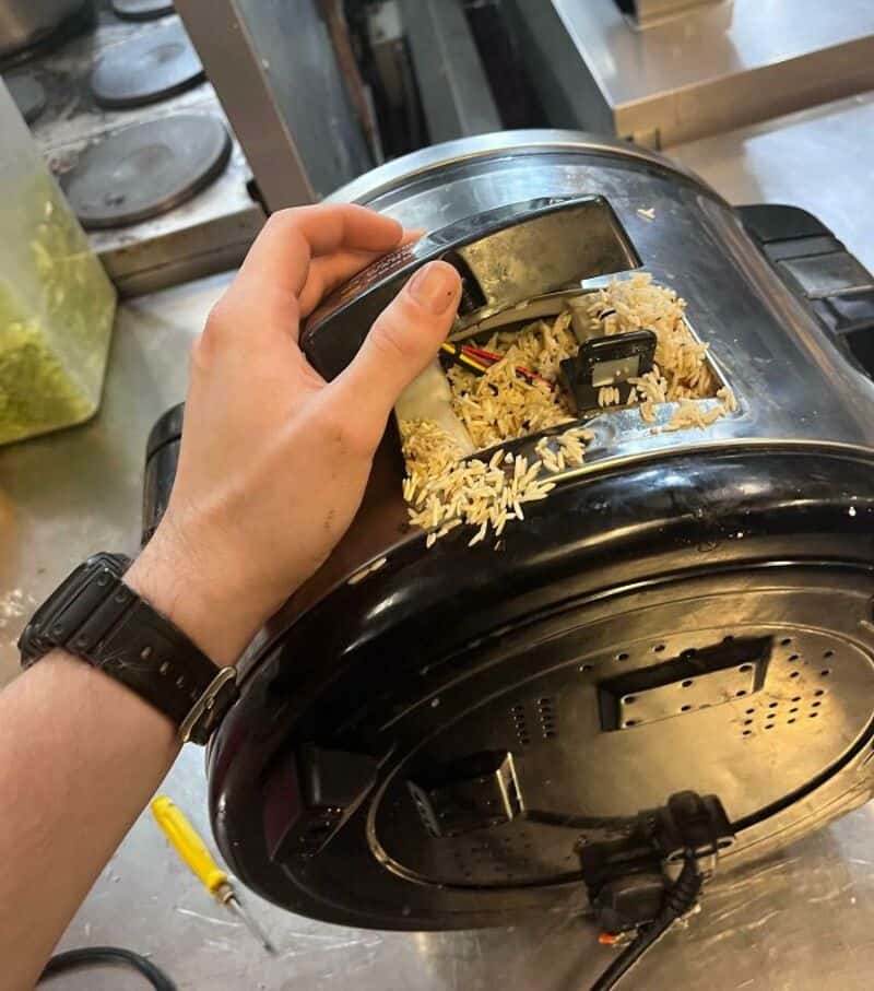 Person's hand opening rice cooker with spilled rice around it.
