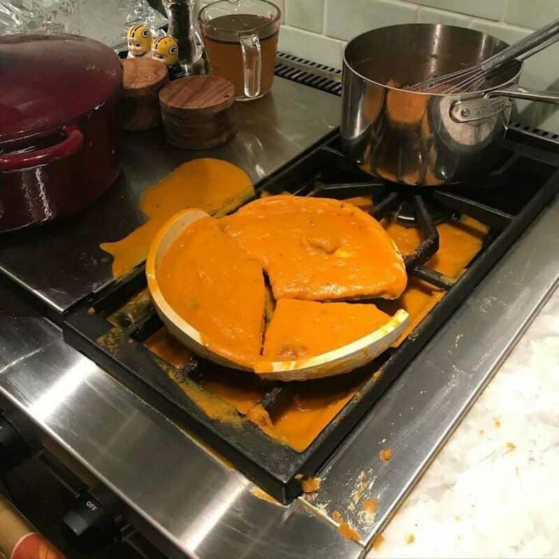 Burnt pumpkin pie on stove with spilled filling and kitchen utensils nearby.