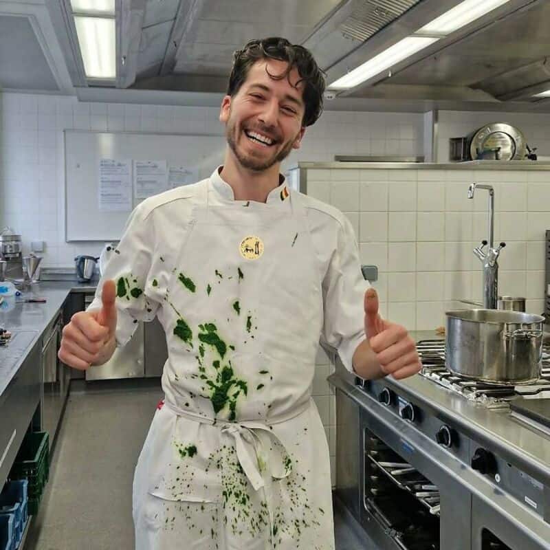 Happy chef with thumbs up in a commercial kitchen, green food stains on uniform.