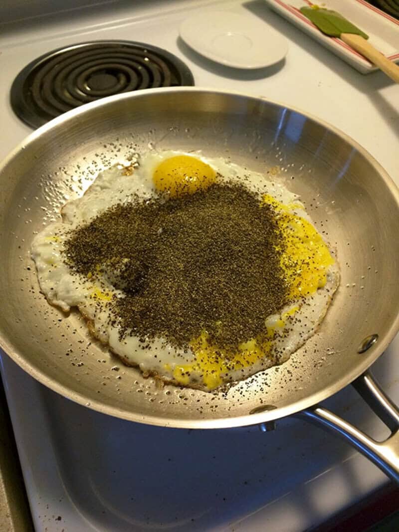 Fried eggs with black pepper in a pan on a stove, with a white plate and utensils nearby.