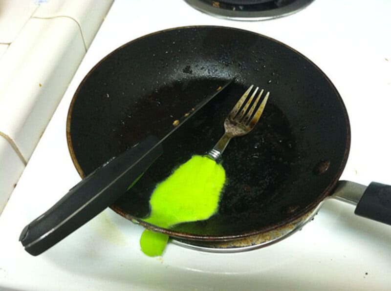 Frying pan with bright green oil, knife and fork inside, on a white stove.