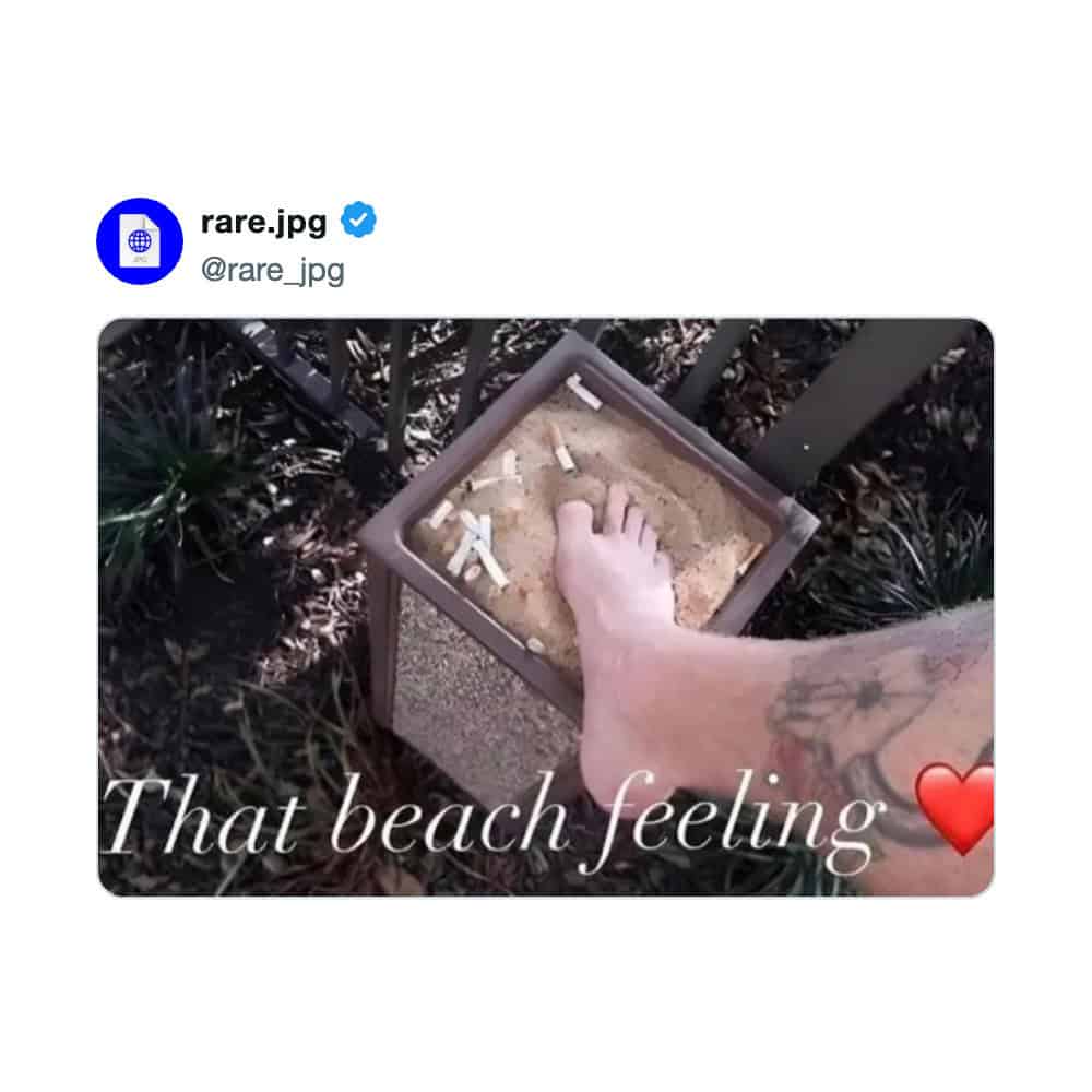 Person's foot in sand-filled box with cigarette butts, captioned 'That beach feeling' with a red ❤️.