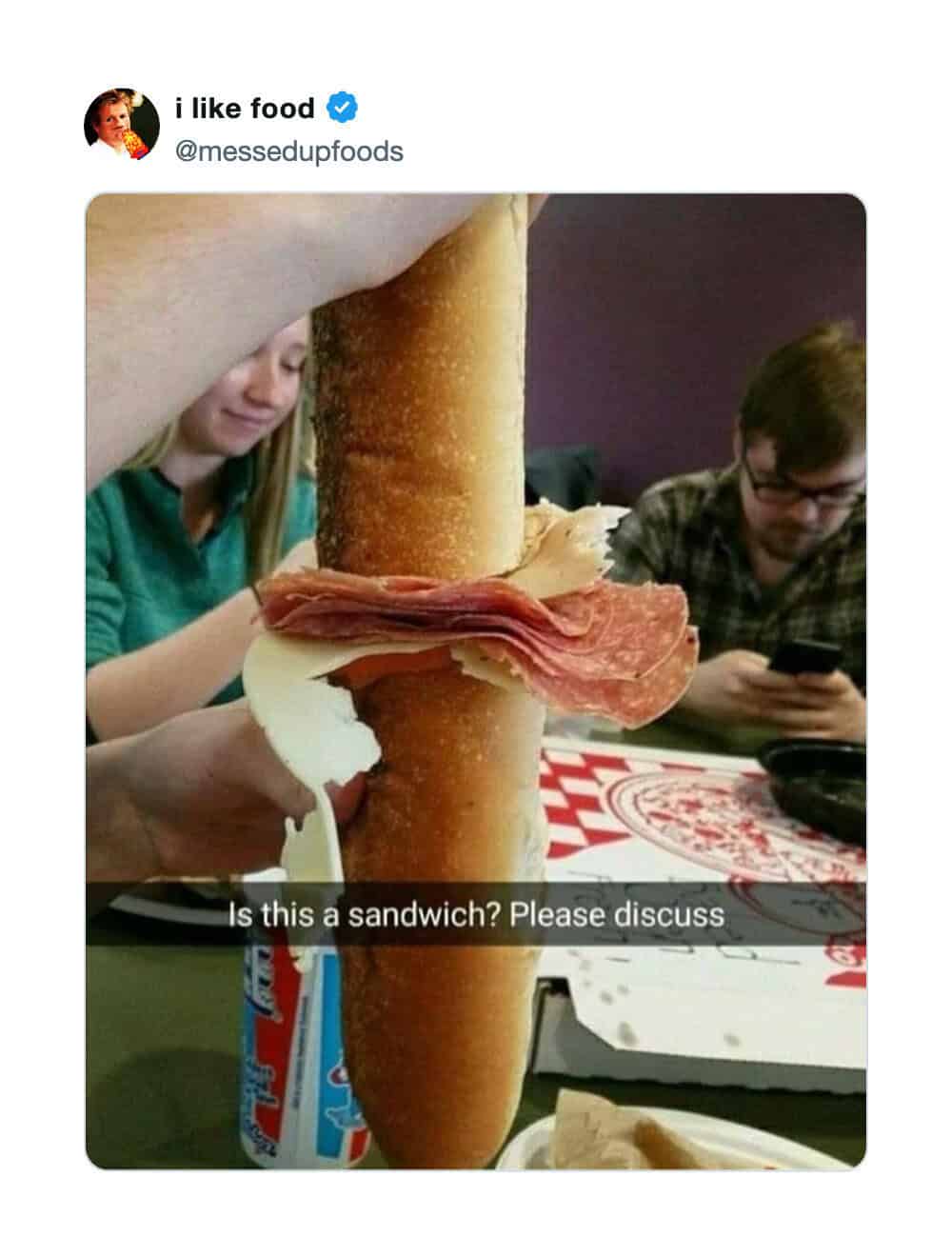 Person holding a sandwich with a large breadstick, with two people in the background.