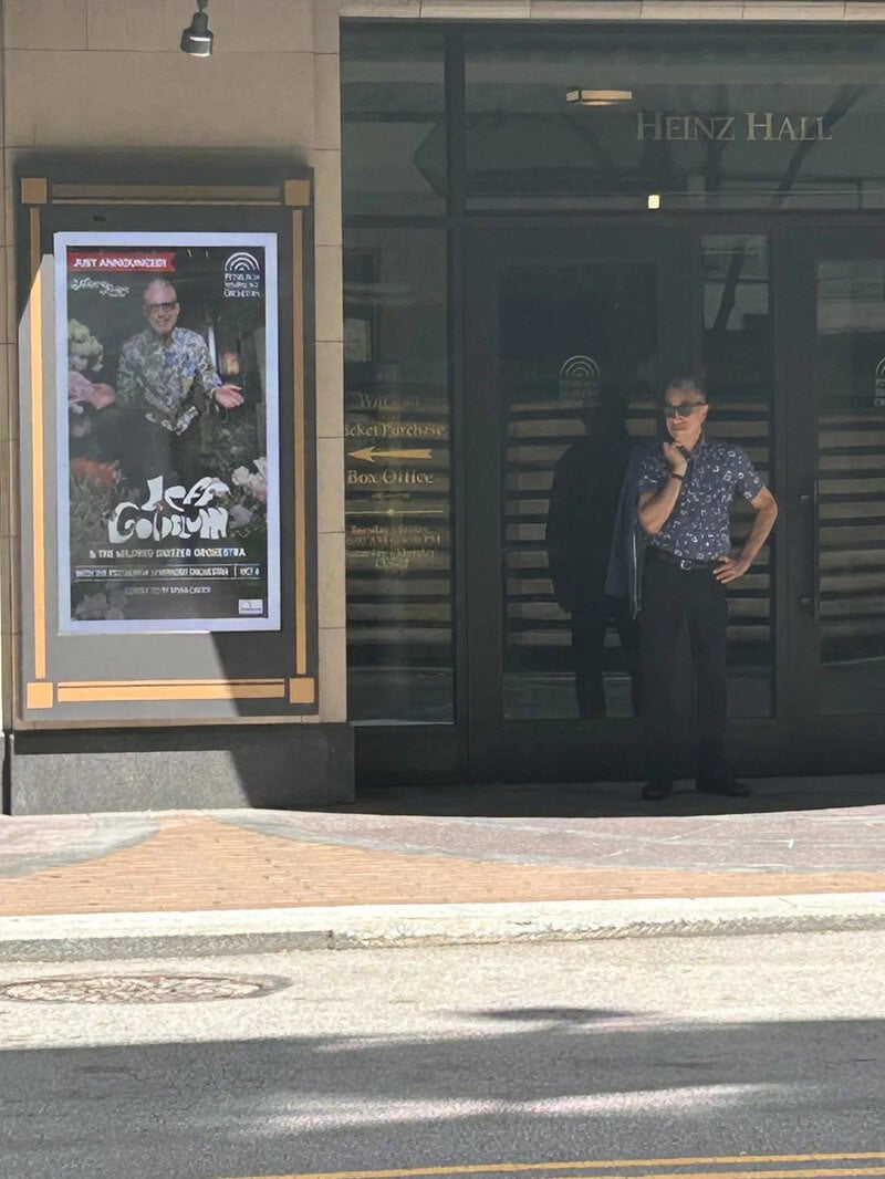 Person standing outside Heinz Hall next to a poster of a man in a floral shirt.