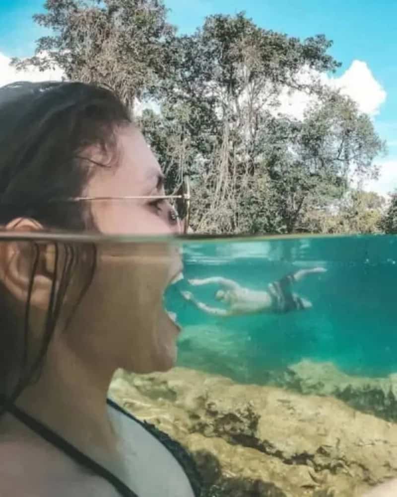 Woman with sunglasses and long hair looking at a fish underwater.