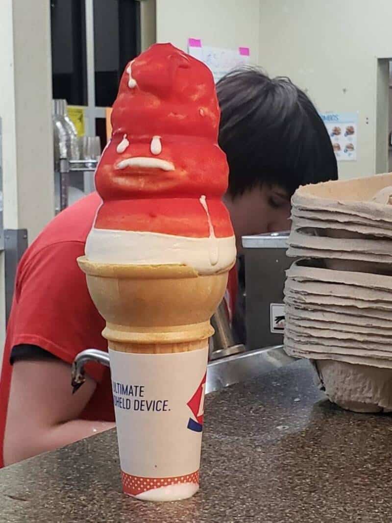 Ice cream cone with red and white ice cream and a sad face, in front of a person and stacks of paper.