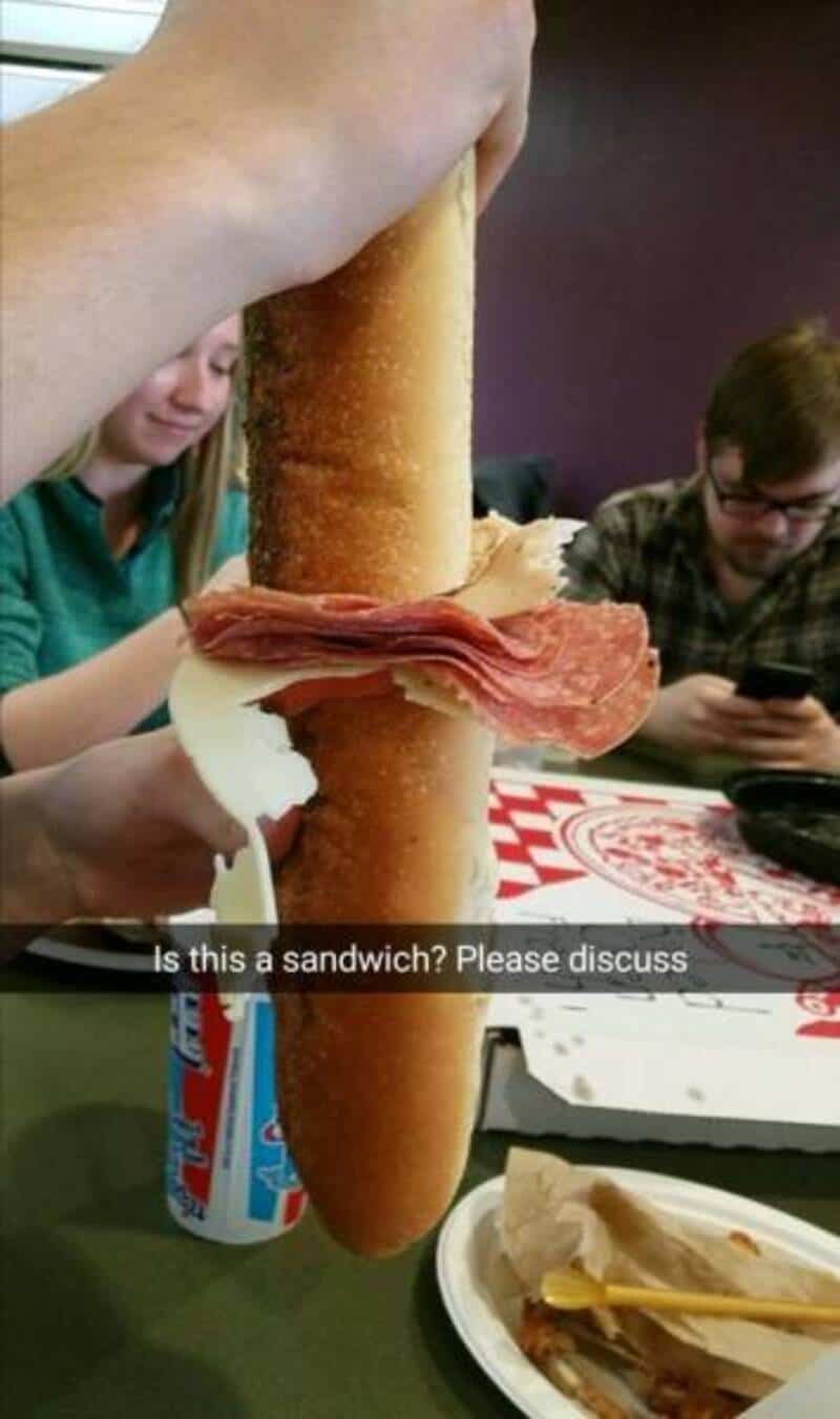 A person holds a large breadstick with ham and cheese, resembling a sandwich.