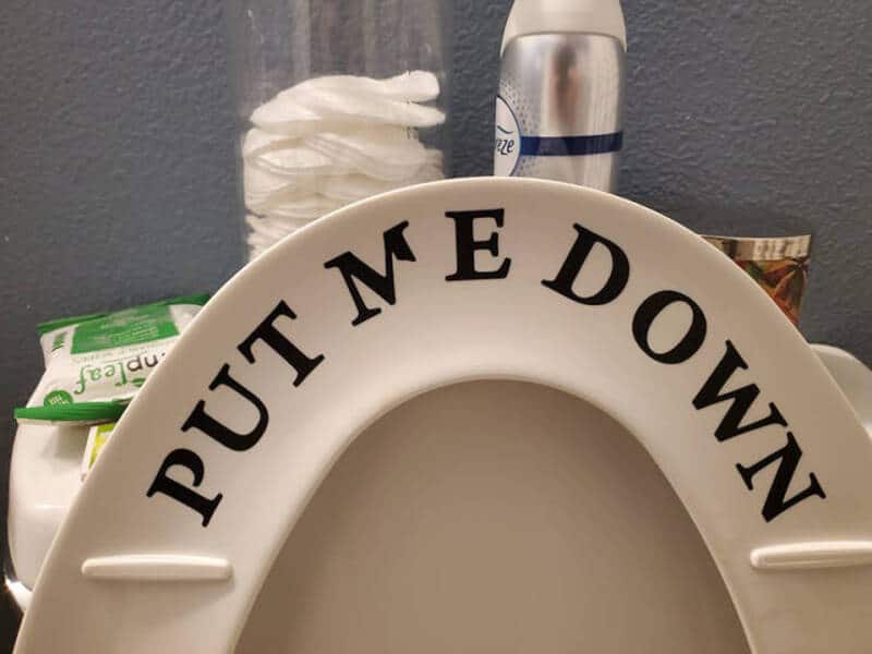 Toilet seat with 'PUT ME DOWN' written on it in black letters.