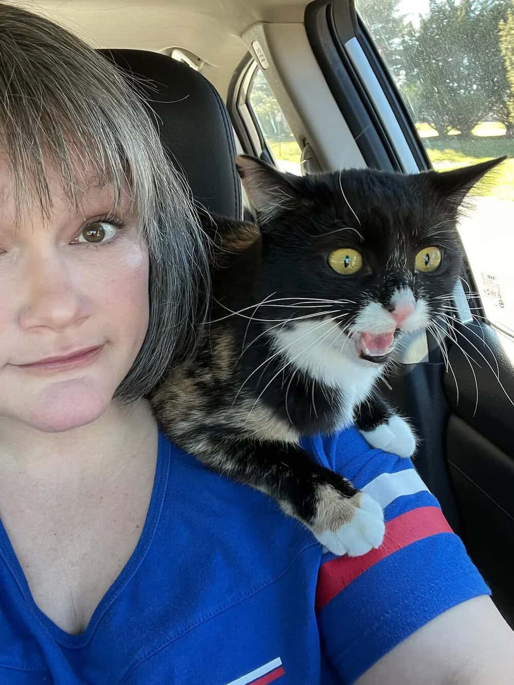 Person with short hair and a black and white cat in a car, both looking at the camera.