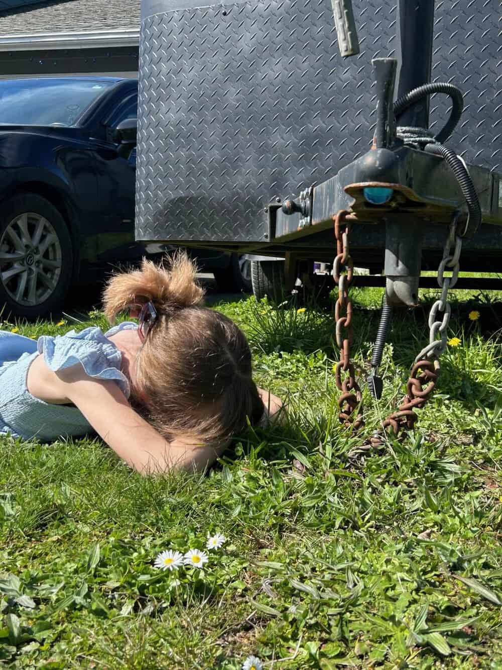 Child lying on grass near trailer hitch, pretending to be trapped.