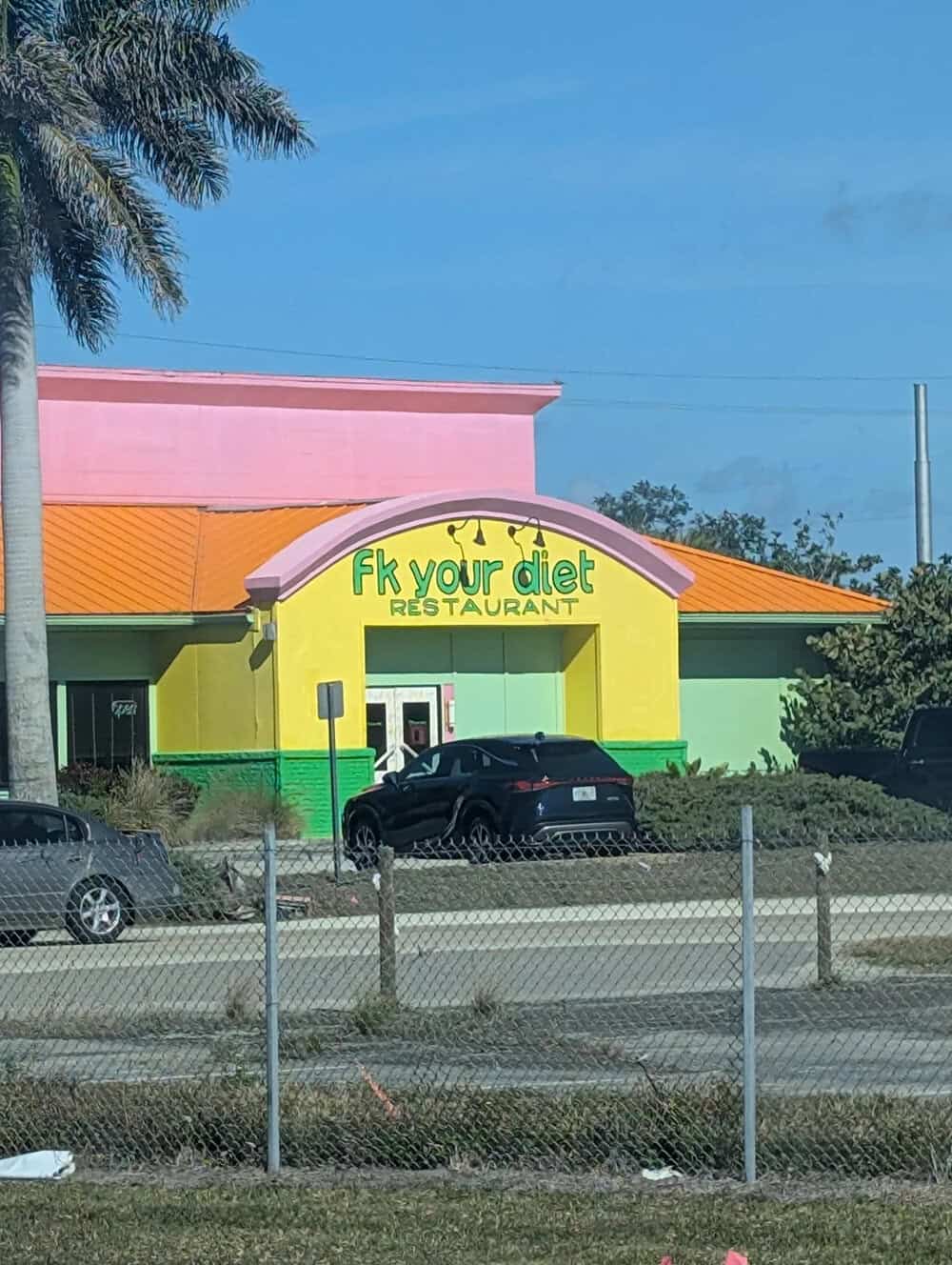 Colorful building with 'FK your diet' sign, cars parked outside, palm tree nearby.