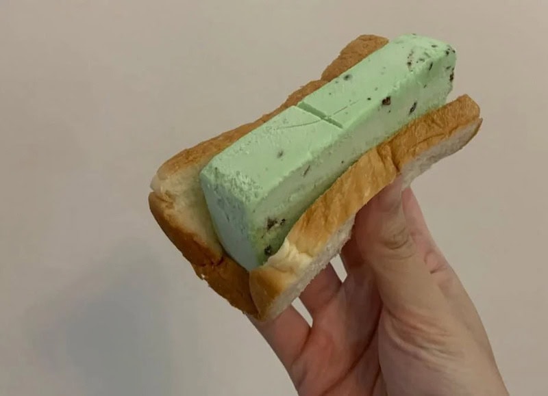 Hand holding a sandwich with green ice cream and bread, against a plain background.