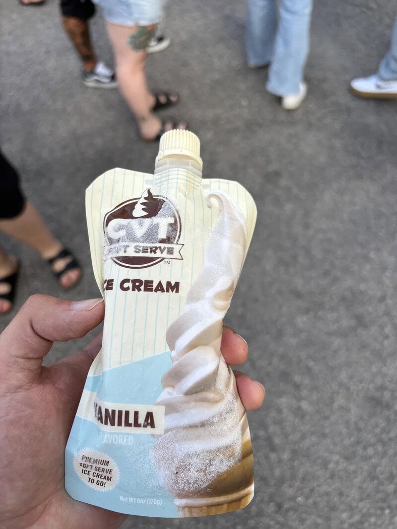 Hand holding vanilla ice cream in a squeeze pouch with people in the background.