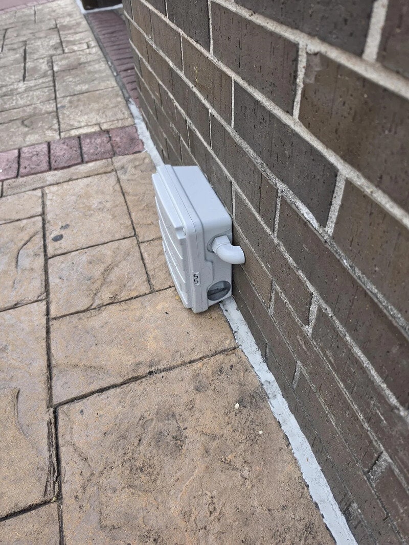 Electrical box on exterior wall with a pipe, next to a brick wall and stone pavement.