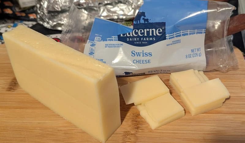Block of Swiss cheese with slices on a wooden surface and cheese package in background.