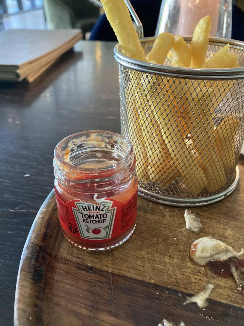 French fries with ketchup on a wooden tray, with a small jar of ketchup and some sauce.