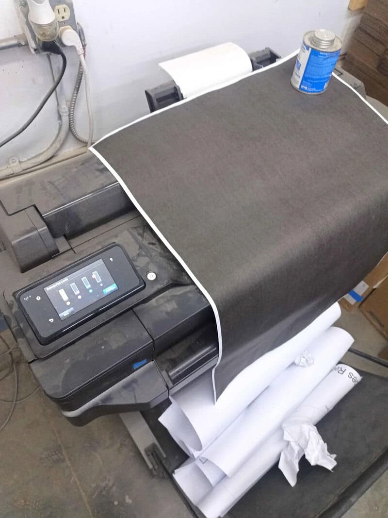 A printer with a large sheet of fabric or paper on it, a can of spray, and rolls of paper underneath.